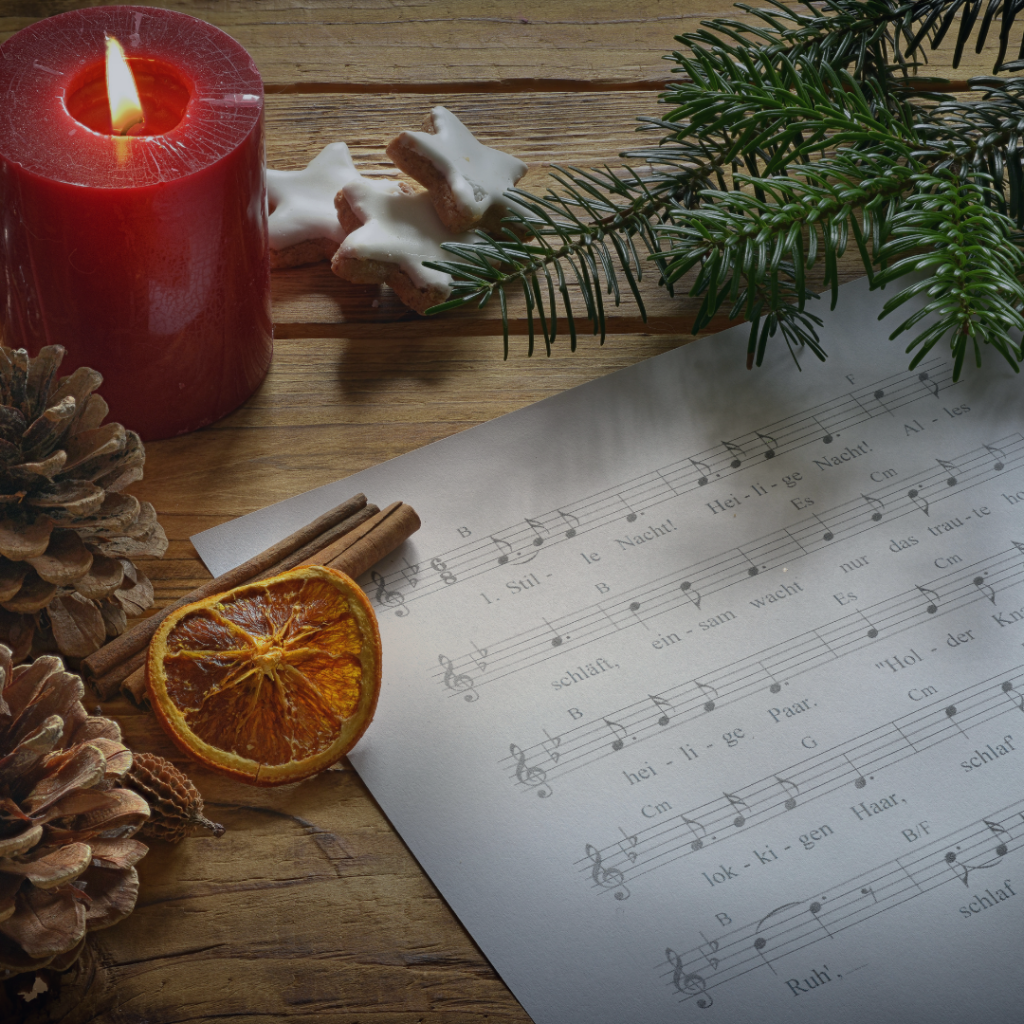 A sheet of music placed on a wooden surface with a red candle, pine cones, cinnamon sticks, candied orange, cookies and a small Christmas tree branch around it